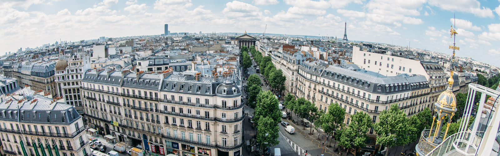 A high-up view of a city with buildings and trees of many different shapes and sizes. The Eiffel Tower is visible in the distance. 